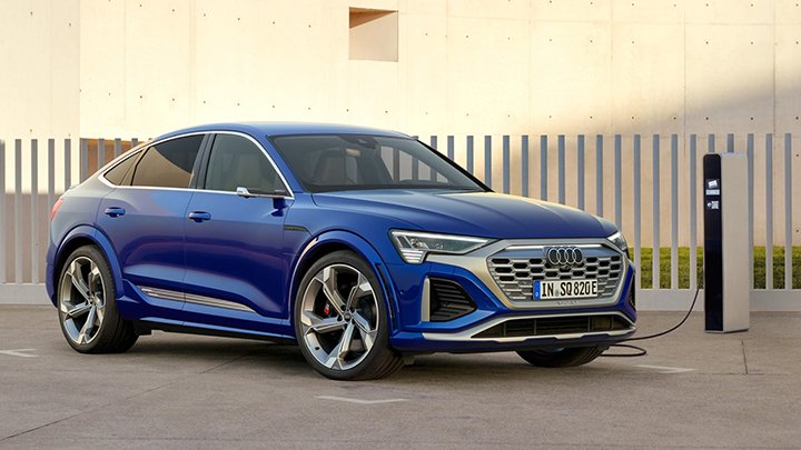 Blue Audi SQ8 Sportback e-Tron parked in front of a small white fence. This EV is plugged into charger.